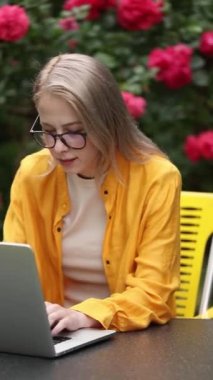 Sarı gömlekli şık bir kadın bahçede dizüstü bilgisayarla çalışıyor ve arka planda güller var.
