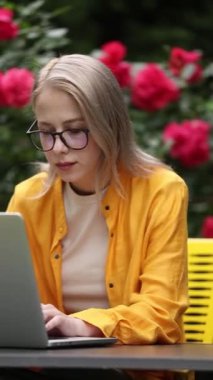 Sarı gömlekli şık bir kadın bahçede dizüstü bilgisayarla çalışıyor ve arka planda güller var.