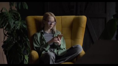 Orta yaşlı beyaz bir kadın, evinde bir koltukta oturmuş, sosyal medyada görüntüler izlerken gülüyor..