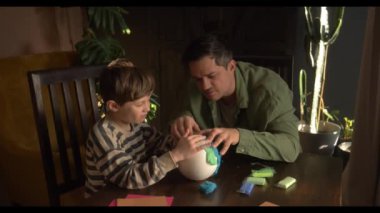 Orta yaşlı Avrupalı bir baba ve okul çağında bir oğul, Dünya Günü 'nü kutlamak için kilden bir Dünya heykeli yapıyorlar, bir masada oturuyorlar, video görüntüleri.