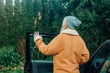 Kahverengi kırbaçlı ceketli ve mavi şapkalı sarışın bir kız. Arka planda çam ağaçları olan bir arabanın kapısının yanında duruyor.