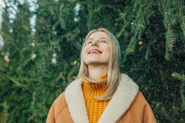 Kar yağarken dışarıda çam ağaçlarının arka planında duran kahverengi kırlangıç ceketli sarışın bir kız.