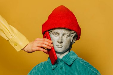 Kırmızı hippi şapkalı ve turkuaz tişörtlü bir gencin antika heykeli sarı arka planda duruyor. Yanında cep telefonu tutan bir kadın var.