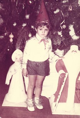 Küçük çocuk 1980 'lerdeki okul yılbaşı partisinde Noel Baba heykelciğinin ve Noel ağacının yanında duruyor.