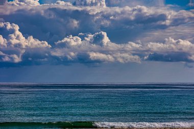 Yumuşak beyaz bulutlar, dingin bir okyanus ufkunun üzerinde geniş mavi bir gökyüzü boyunca uzanıyor ve sakin bir manzara yaratıyor. Suyun üzerindeki bulutların yansıması huzurlu ortamı artırır..