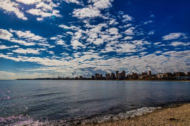 Alicante kıyı silueti tüylü bulutlarla dolu dramatik bir gökyüzü altında uzanıyor. Gökdelenler uzak kıyı boyunca uzanırken, yumuşak dalgalar kayalık bir plaja çarpıyor..