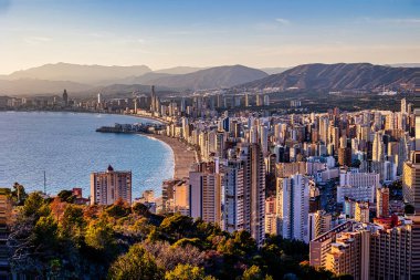 Bir kıyı şehri Benidorm, kıyı şeridi boyunca yığılan yüksek binalarla batan güneşin sıcacık parıltısıyla yıkanır.