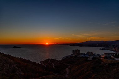 Göz kamaştırıcı bir günbatımı, kıyı şehrine sıcacık bir ışıltı saçar. Güneş sakin denizi güzel bir şekilde yansıtır.