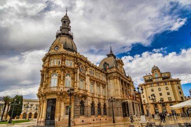 Süslü, tarihi bir bina parçalı bulutlu bir gökyüzünün altında görkemli bir şekilde duruyor. Karmaşık mimari detaylar ve belirgin bir saat kulesi var.