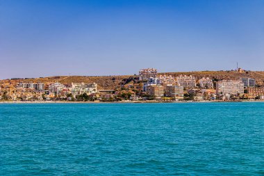 Turkuvaz suları üzerinde kıyı şehri manzarası görülebiliyor. Sahili kayan tepelere karşı dizen sayısız bina var.