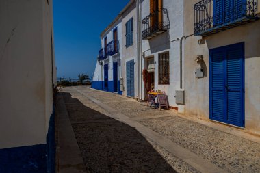 Dar bir kaldırım taşı caddesi açık, güneşli bir gökyüzünün altında parlak mavi panjurları ve kapıları olan binalarla çevrili..
