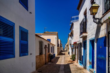 Dar bir kaldırım taşı caddesi açık, güneşli bir gökyüzünün altında parlak mavi panjurları ve kapıları olan binalarla çevrili..