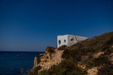 Derin mavi denize bakan kayalık bir uçurumun üzerinde küçük beyaz bir bina duruyor..
