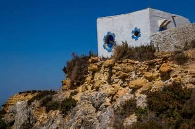 Derin mavi denize bakan kayalık bir uçurumun üzerinde küçük beyaz bir bina duruyor..