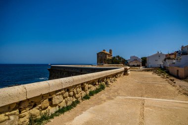 Sağlam bir duvarı olan taş bir patika okyanusa bakıyor. Beyaz binaları olan küçük bir sahil köyüne doğru gidiyor.