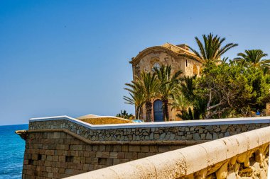 Tarihi bir taş bina sahil boyunca açık mavi gökyüzünün altında yemyeşil palmiye ağaçları ile çevrilidir.