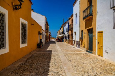 Güneşli bir kaldırım taşı caddesi renkli balkonlu ve pencereli binalarla çevrili, canlı ve davetkar bir atmosfer yaratıyor.
