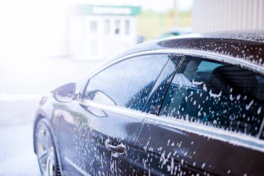 Koyu renkli bir arabanın vücuduna parlak güneş ışığında beyaz köpük sürülür.