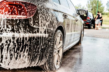 Gündüz vakti araba yıkamada koyu renk bir arabanın gövdesine beyaz köpük sürülür.