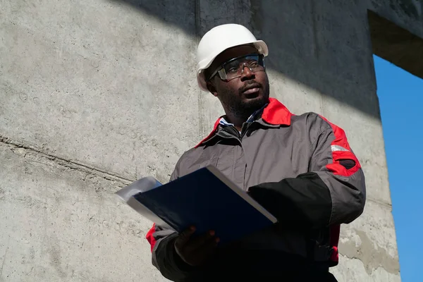 African american worker stands at construction site with work papers and looks around