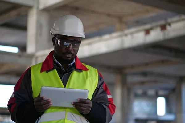 African american worker in hard hat and protective goggles stands at construction site, holds in hands tablet computer and looks at the camera