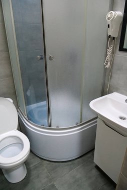 Toilet room interior with white toilet bowl with open lid, shower cabin, wash basin, hairdryer, cabinet under the washbasin and grey wall and floor tiles