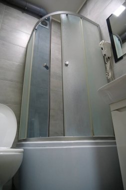 Tuvaletin içi açık kapaklı, duş kabini, lavabo, saç kurutma makinesi, lavabo ve gri duvarın altındaki dolap ve yerdeki fayanslarla dolu.