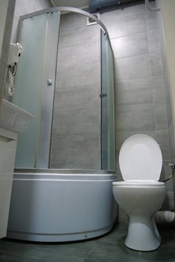 Toilet room interior with white toilet bowl with open lid, shower cabin, wash basin, hairdryer, cabinet under the washbasin and grey wall and floor tiles