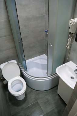 Toilet room interior with white toilet bowl with open lid, shower cabin, wash basin, hairdryer, cabinet under the washbasin and grey wall and floor tiles