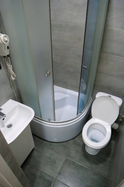 Toilet room interior with white toilet bowl with open lid, shower cabin, wash basin, hairdryer, cabinet under the washbasin and grey wall and floor tiles