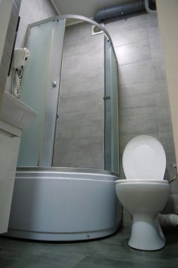 Tuvaletin içi açık kapaklı, duş kabini, lavabo, saç kurutma makinesi, lavabo ve gri duvarın altındaki dolap ve yerdeki fayanslarla dolu.
