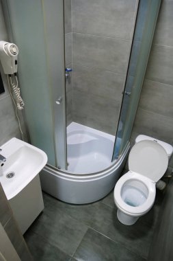 Toilet room interior with white toilet bowl with open lid, shower cabin, wash basin, hairdryer, cabinet under the washbasin and grey wall and floor tiles
