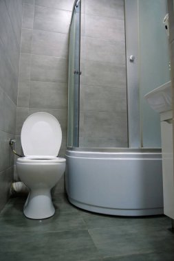 Toilet room interior with white toilet bowl with open lid, shower cabin, wash basin, hairdryer, cabinet under the washbasin and grey wall and floor tiles