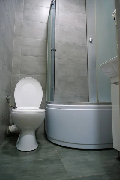 stock image Toilet room interior with white toilet bowl with open lid, shower cabin, wash basin, hairdryer, cabinet under the washbasin and grey wall and floor tiles