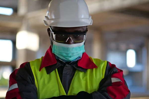 stock image African american builder in medical protective face mask stands at construction site and looks at the camera, close-up shot