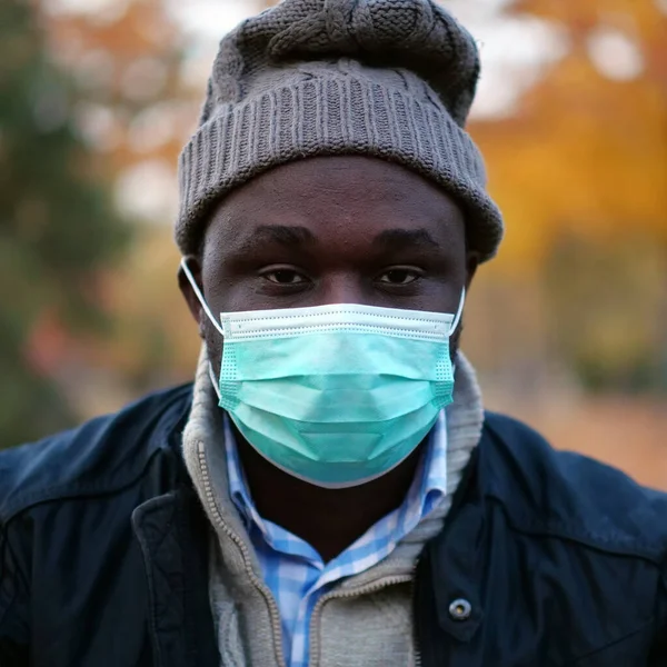 Stock image African american man in medical protective face mask and winter hat sits in autumn city park and looks at camera