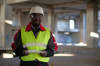 Siyah şapkalı ve koruyucu gözlüklü Afro-Amerikan işçiler inşaat alanında duruyor, tablet bilgisayarı tutuyor ve kameraya bakıyor.