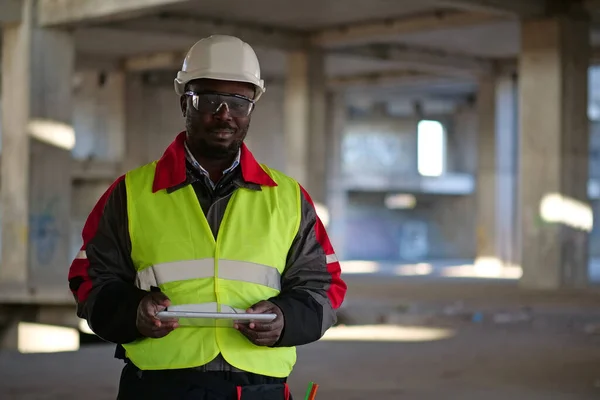 African american worker in hard hat and protective goggles stands at construction site, holds in hands tablet computer and looks at the camera