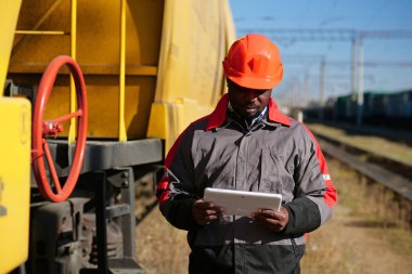 Inspector of wagons at freight train station looks in tablet computer. African american railway man with tablet pc at freight train terminal