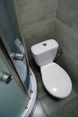 Toilet room interior with white toilet bowl, shower cabin and grey wall tiles