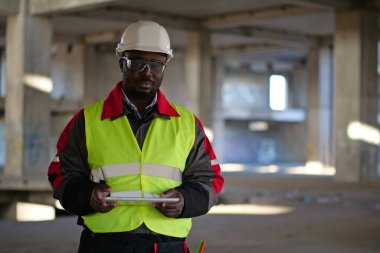 Siyah şapkalı ve koruyucu gözlüklü Afro-Amerikan işçiler inşaat alanında duruyor, tablet bilgisayarı tutuyor ve kameraya bakıyor.