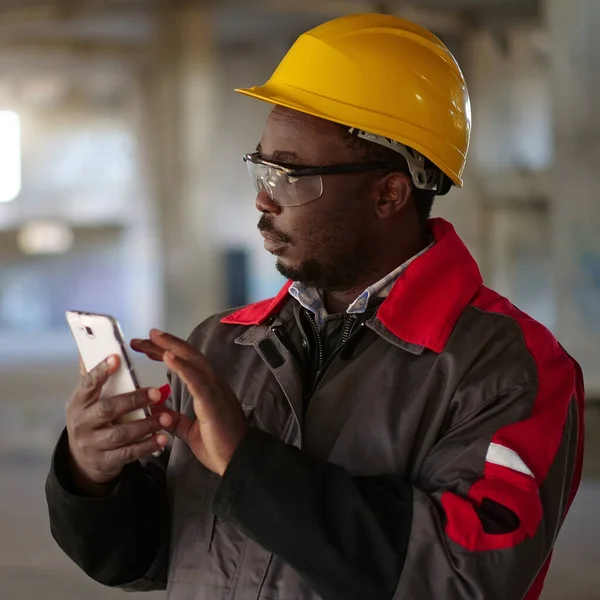 stock image African american workman in yellow hard hat with smartphone stands at construction site. Worker holds in hands smartphone