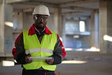 Siyah şapkalı ve koruyucu gözlüklü Afro-Amerikan işçiler inşaat alanında duruyor, tablet bilgisayarı tutuyor ve kameraya bakıyor.