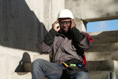 İnşaat alanındaki merdivenlerde oturan Afrikalı Amerikalı işçiler. Kendini iyi hissetmiyor ve başı ağrıyor. Kafasına dokunuyor.