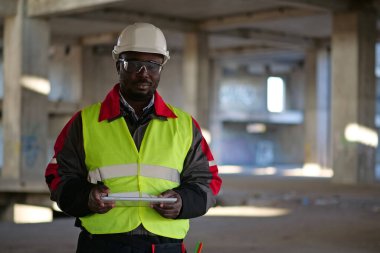 Siyah şapkalı ve koruyucu gözlüklü Afro-Amerikan işçiler inşaat alanında duruyor, tablet bilgisayarı tutuyor ve kameraya bakıyor.