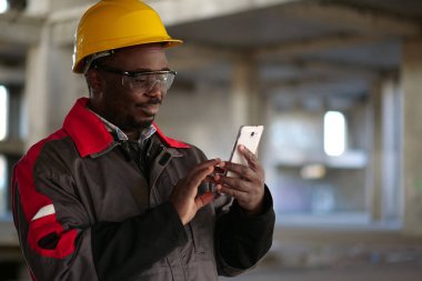 İnşaat alanında sarı şapkalı, akıllı telefonlu Afro-Amerikan işçiler var. İşçi el ele tutuşur, akıllı telefon ve gülümser.