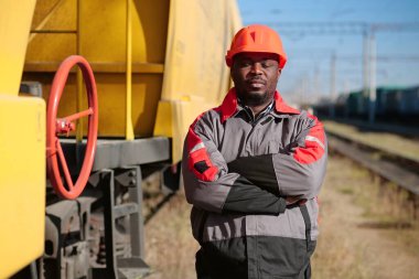 Afrikalı Amerikan demiryolu işçisi, yük treni terminalinde gözleri kapalı duruyor. Demiryolu işçisi üniformalı ve kırmızı şapkalı, kameraya bak.