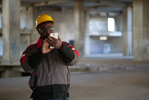スマートフォンを備えた黄色のハード帽子のアフリカ系アメリカ人労働者は 建設現場に立っています スマートフォンを手に持つ労働者 — ストック写真
