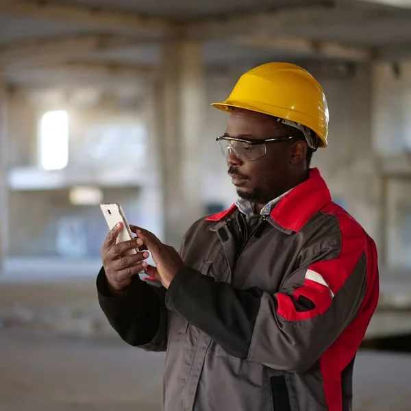 スマートフォンを備えた黄色のハード帽子のアフリカ系アメリカ人労働者は 建設現場に立っています スマートフォンを手に持つ労働者 ストック画像