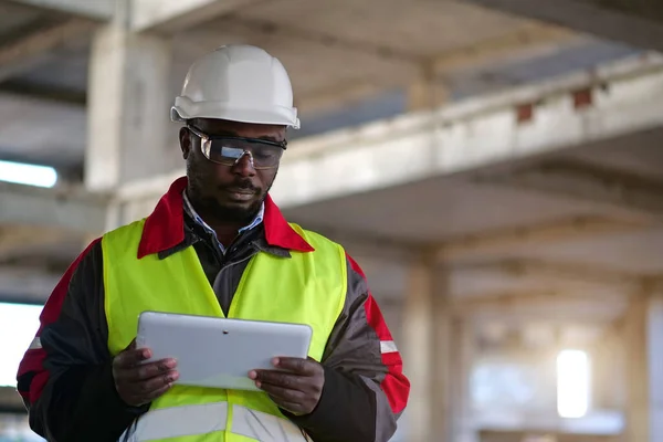 African american worker in hard hat and protective goggles with closed eyes stands at construction site, holds in hands Tablet PC